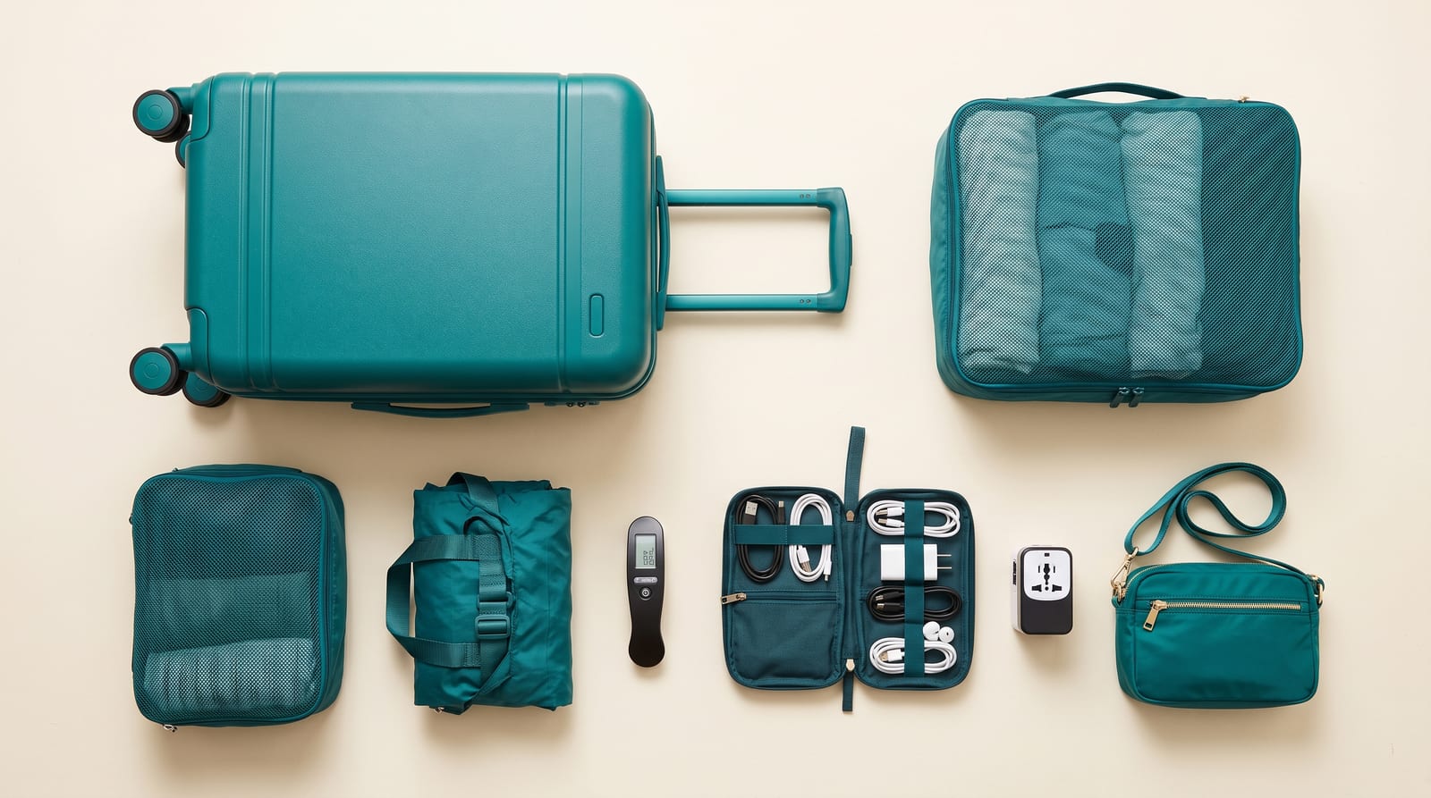 Teal travel gear set on a cream background — hardside carry-on suitcase, packing cubes, foldable duffel, luggage scale, cable organizer with chargers, universal adapter, and a crossbody bag